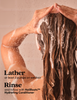 A person with long, wet brown hair lathered in foam, shown from the back while washing. Text on the left reads, 'Lather at least 3 pumps on wet hair Rinse and follow with HoliRoots™ Hydrating Conditioner.