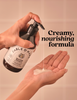 A hand holds a pump bottle of Fable & Mine Hydroscopic Shampoo above another hand, with creamy shampoo being dispensed into the palm. The background is a soft beige tiled wall, and bold text on the right reads 'Creamy, nourishing formula'.